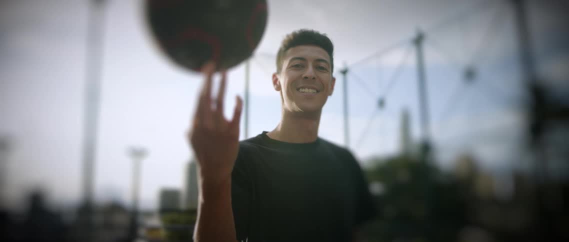 A young man wearing a black shirt smiles brightly at the camera while spinning a darkcolored ball on his right index finger in an outdoor sports court