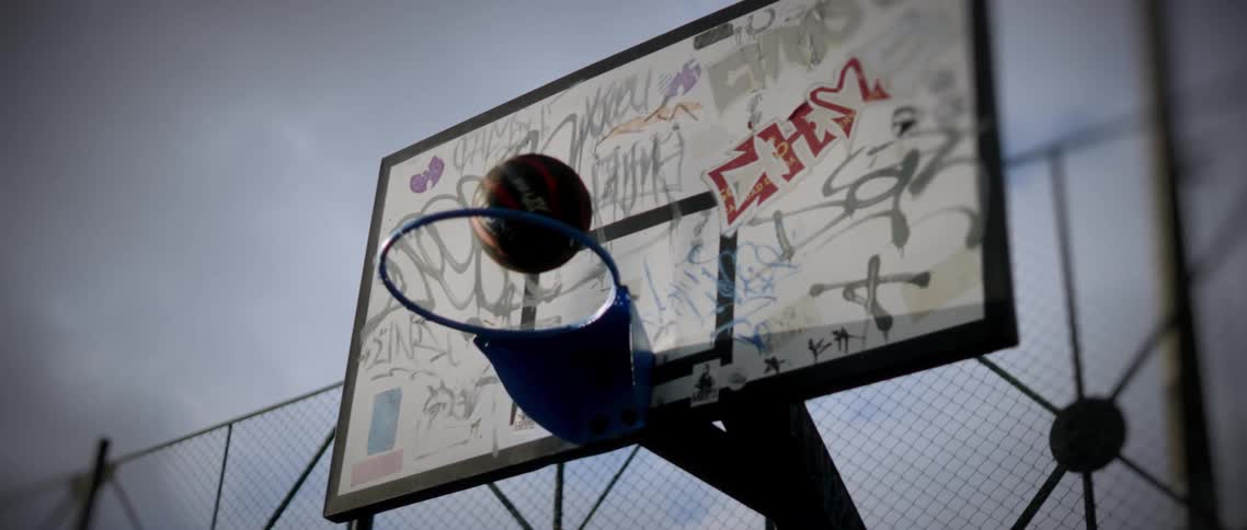 A lowangle shot captures an outdoor basketball backboard heavily covered in gray and white graffiti featuring a bright blue rim as a darkcolored basketball flies up and successfully drops through the hoop under an overcast sky