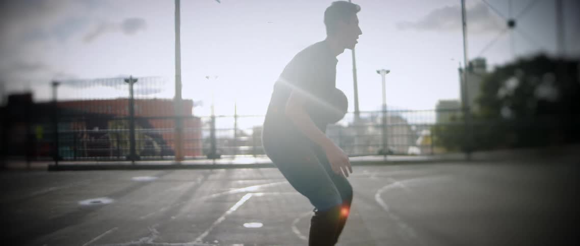 This video shows a young male in a dark tshirt and pants dribbling a basketball on an outdoor courtthe footage is shot against bright sunlight creating a highcontrast silhouette effect and the watermark filmsupply is visible across the middle of