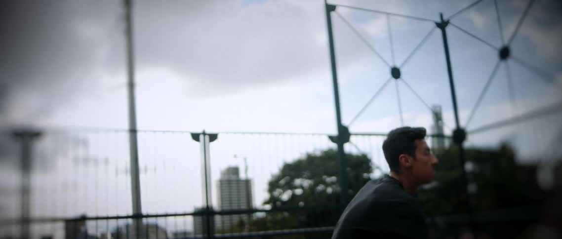 A young man in a black shirt is shown practicing basketball outdoors on a cloudy day he dribbles the black and orange ball close to the ground then swiftly moves to shoot releasing the ball toward a graffiticovered backboard and hoop