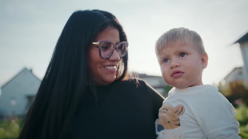 Smiling warmly a woman holds a toddler and waves enthusiastically at the camera briefly turning to speak to the child as bright sunlight flares around them in a quiet neighborhood