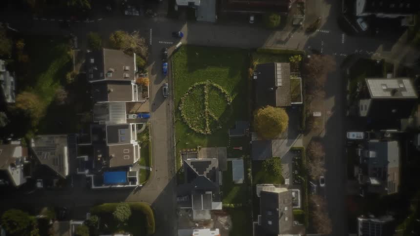 A direct topdown aerial view slowly descends over a suburban neighborhood street grid focusing on a large central green lawn where a prominent peace sign symbol has been cut into the grass