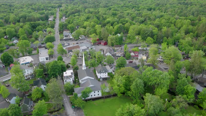 A church with a tall steeple is at the center of a very small town neighborhood dotted with older working class homes and surrounded by woodlands