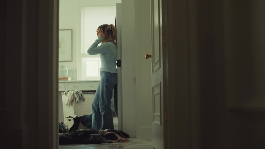 A depressed woman sits on the laundry room floor seen through a doorway