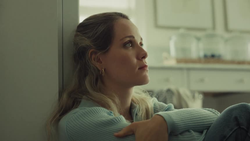The video shows a closeup of a blonde woman sitting on the floor leaning against a wall in what appears to be a brightly lit kitchen or living area she is wearing a light blue ribbed sweater and jeansshe looks thoughtful and slightly wistful gazin