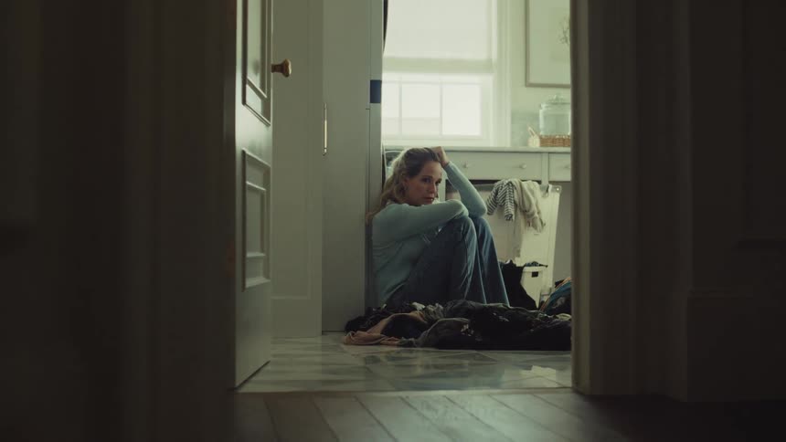 A young woman sits on the floor inside her laundry room appearing sad and lost in thought