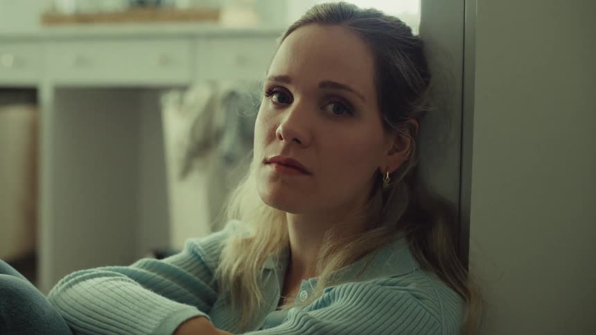 Leaning against a wall in a softly lit room a young woman gazes directly at the camera with a somber contemplative expression blinking slowly as she sits in silence