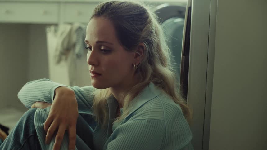 A woman is sitting on the floor of a laundry room leaning against a surface with her arms wrapped around her raised knee she alternates between looking pensively down and looking up and away with a troubled expression suggesting deep thought or sadnes