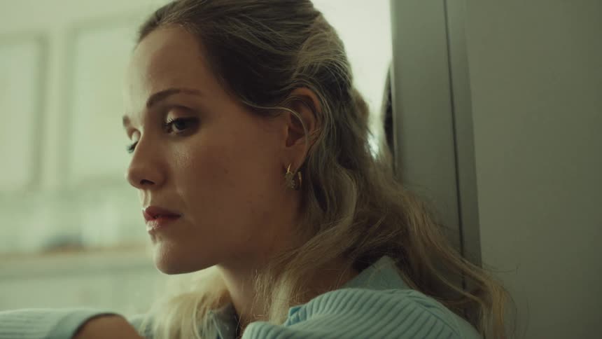 A young woman with long blonde hair and wearing a light blue ribbed sweater is leaning against a wall or cabinet looking distressed she is seen from the side looking down or slightly away for the first half of the clip before lifting her gaze upwards