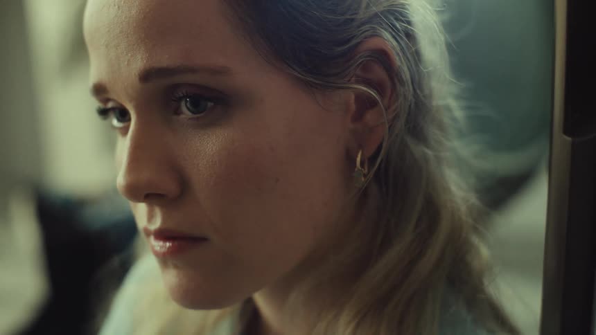A closeup shot focuses on the side profile of a young woman with long blonde hair and a gold hoop earring she initially looks down with a pensive expression then slowly raises her eyes to look forward