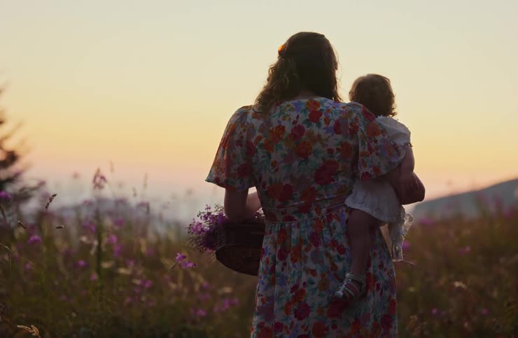A young woman stands in a garden at sunset carrying a baby in her arms while holding a flower plant