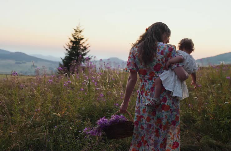 Woman carrying a child and a basket with purple flowers in a field at sunset