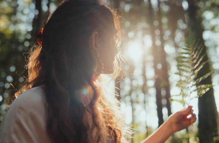 Woman holding a fern leaf while looking at sunlight filtering through the forest