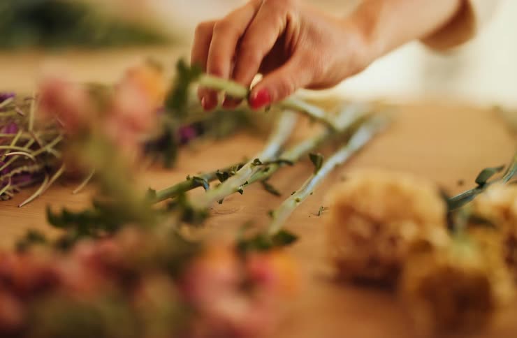 A closeup shot shows a persons hand with red nail polish delicately picking up green flower stems scattered on a wooden table surrounded by other fresh and dried flowers