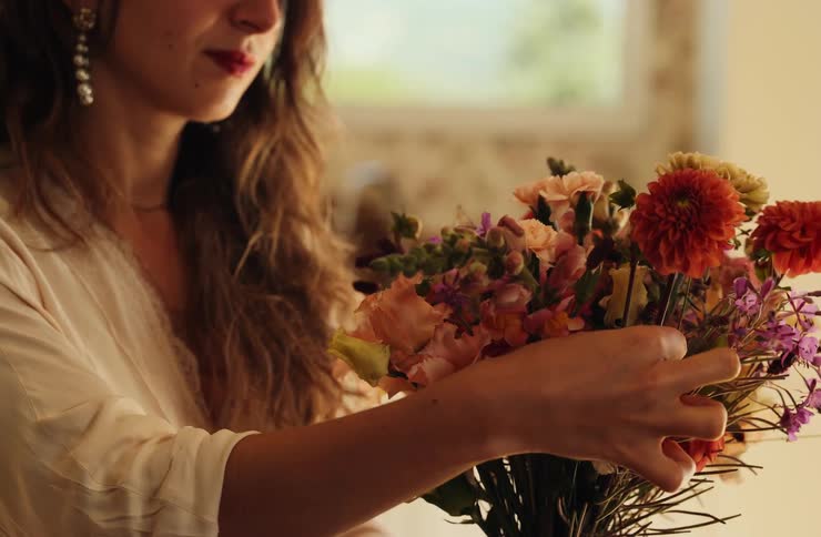 A young woman holds a flower bouquet while sorting through flower plants indoors at midday