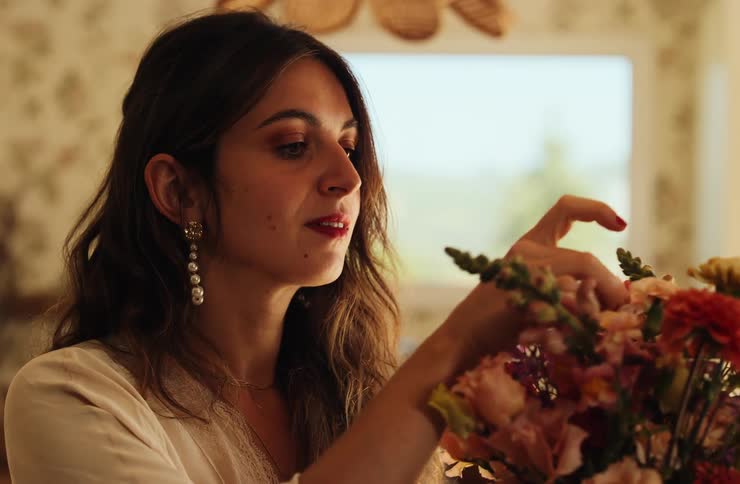 A beautiful woman with long dark hair and drop pearl earrings arranges a colorful bouquet of flowers indoors in warm natural light