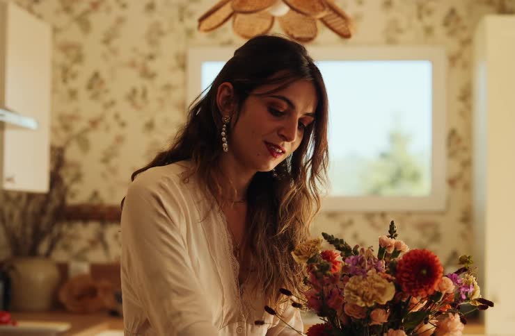 Woman arranges flowers in a home setting with floral wallpaper and window in the background