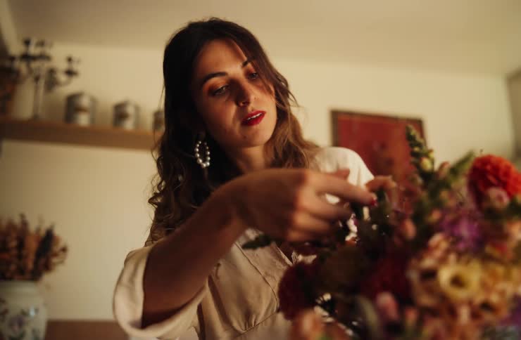 Woman examines floral arrangement in front of shelves with jars and decorative frame