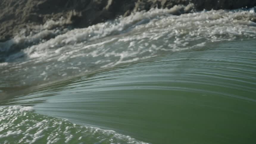 Smooth wave in foreground contrasts with foamy turbulence near rocky shore in the background