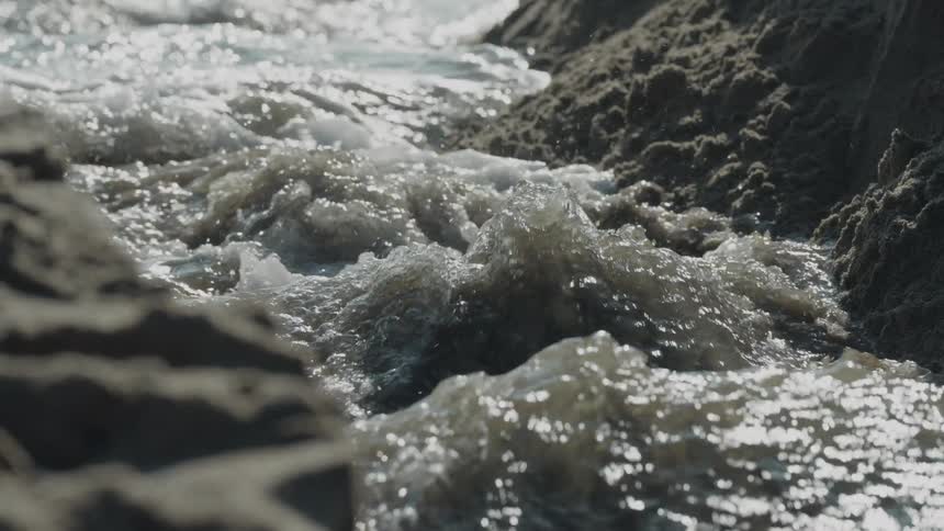 Water flows between sand formations with sunlight reflections and foam in the background