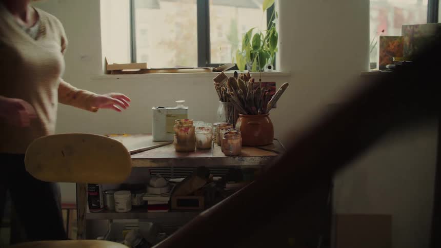 A person organizes paint brushes and jars on a cluttered sunlit studio workbench
