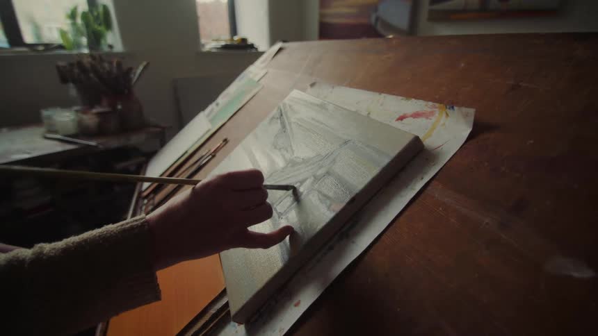 An older woman paints on an inclined surface in a sunlit art filled room