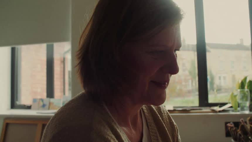 The video is a medium closeup shot of an older womans profile illuminated by soft light coming from a large window behind hershe has short reddishbrown hair and a pleasant subtle smile on her face suggesting she is listening to or engaging with