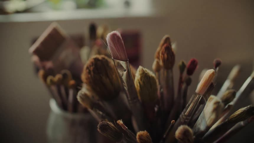 A closeup shot focuses on a dense cluster of art brushes of various sizes and colors held upright in a container with soft natural light