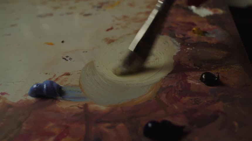 A closeup shot focuses on a paintbrush actively swirling thick white paint on a wooden artists palette to mix colors leaving a noticeable circular stroke pattern