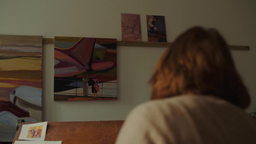 Woman observes wall with colorful paintings and smaller artworks on shelf in creative studio