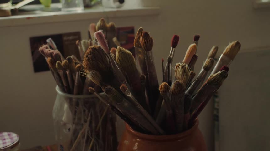 Two containers one glass and one terracottacolored ceramic densely packed with varioussized art brushes their paintstained bristles catching the dim light coming from a window in the background