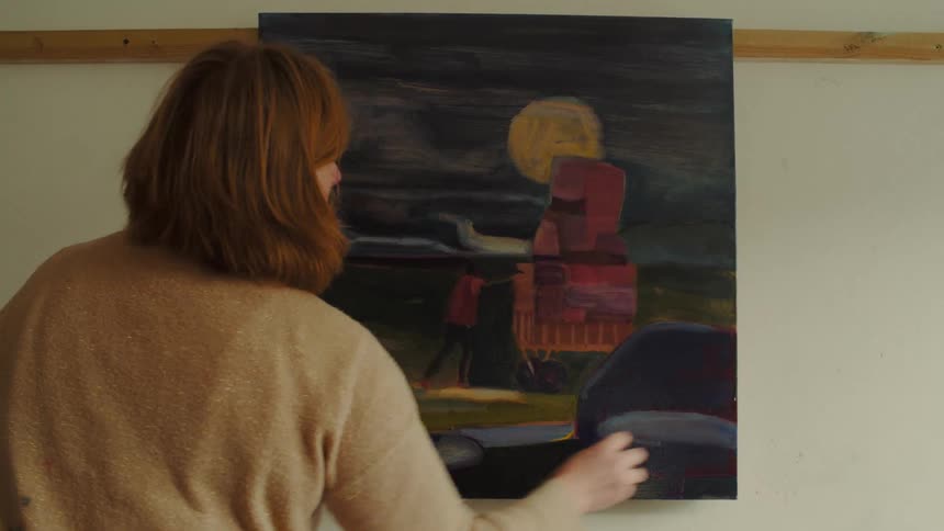 Woman with reddishbrown hair adjusts abstract painting with colorful shapes on white wall