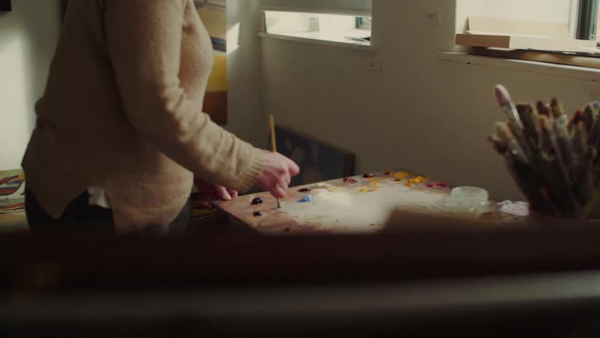 A woman wearing a beige sweater is shown in a studio environment intently using a paintbrush to mix various oil colors on a wooden palette before briefly pausing to wipe her face with several completed abstract canvases hanging on the wall behind her
