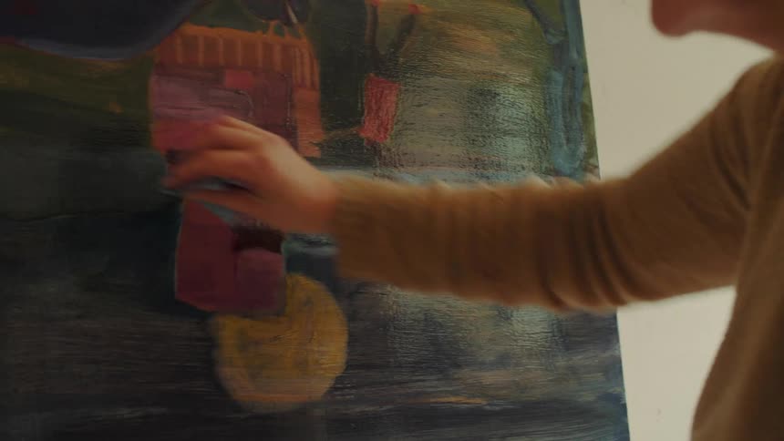An artist viewed from behind works on a large canvas featuring bold abstract shapes and colors using a rag to blend and apply paint to the dark background area