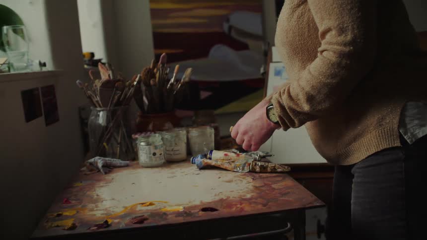Paint tube being handled over a painting table filled with brushes and art supplies