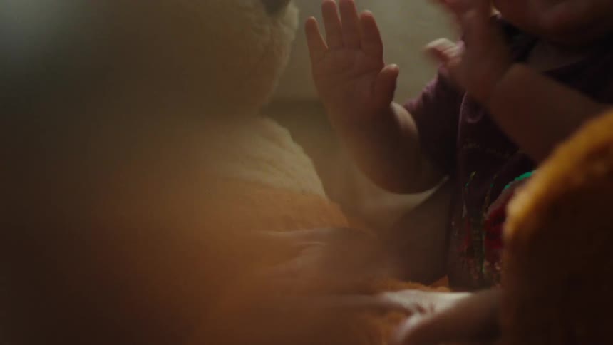 A baby in a persons arms slaps on a large formation of dough on a table in a kitchen
