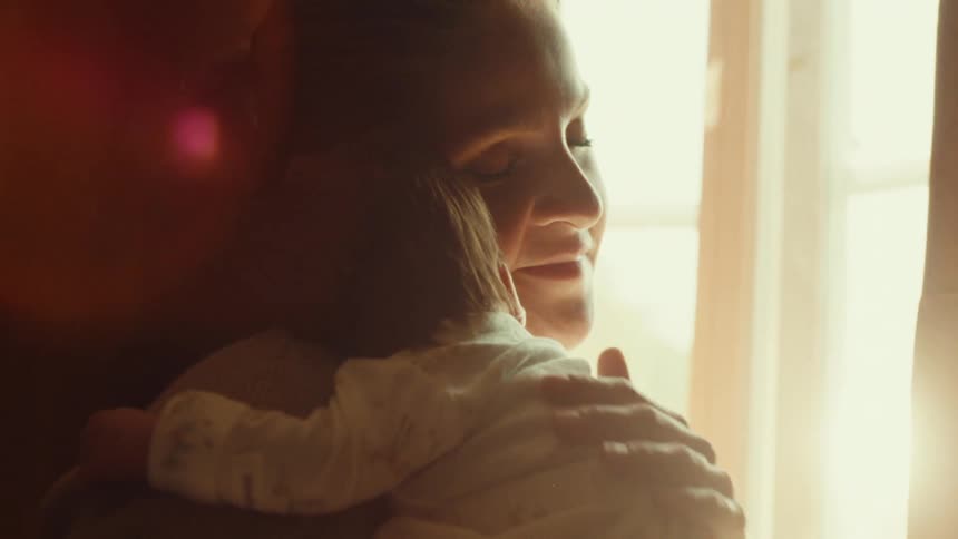 A mother holds her baby rubbing its back in front of a window