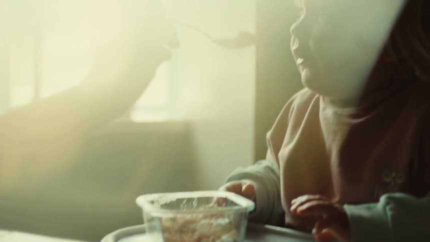 A young child sits at a table reaching for a small clear container of food possibly oatmeal or cereal while an adults hand feeds them with a spoon the scene is heavily backlit by bright sunlight streaming through a window creating a warm hazy and