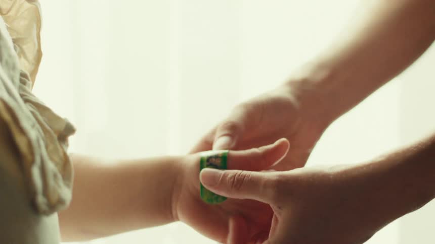 An adults hands gently wrapping a patterned green bandage around the injured finger of a childs hand with a brightly backlit of an indoor setting
