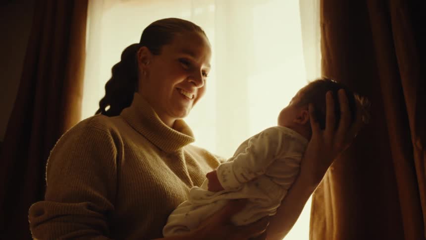 New mother happily kissing her newborn standing by a window