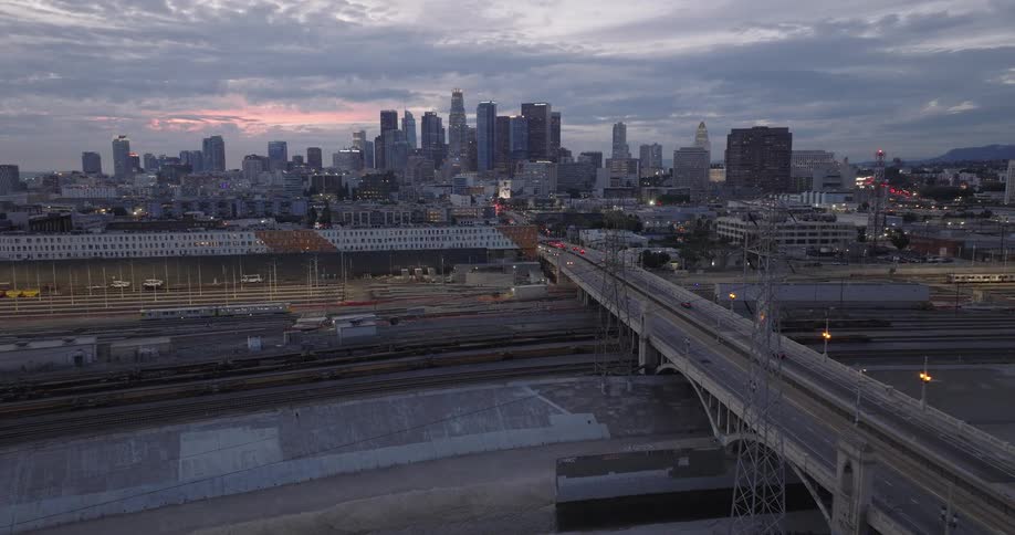 An aerial wide shot captures a sprawling downtown Los Angeles at twilight featuring a large bridge with passing traffic spanning over a concrete river channel and railyard