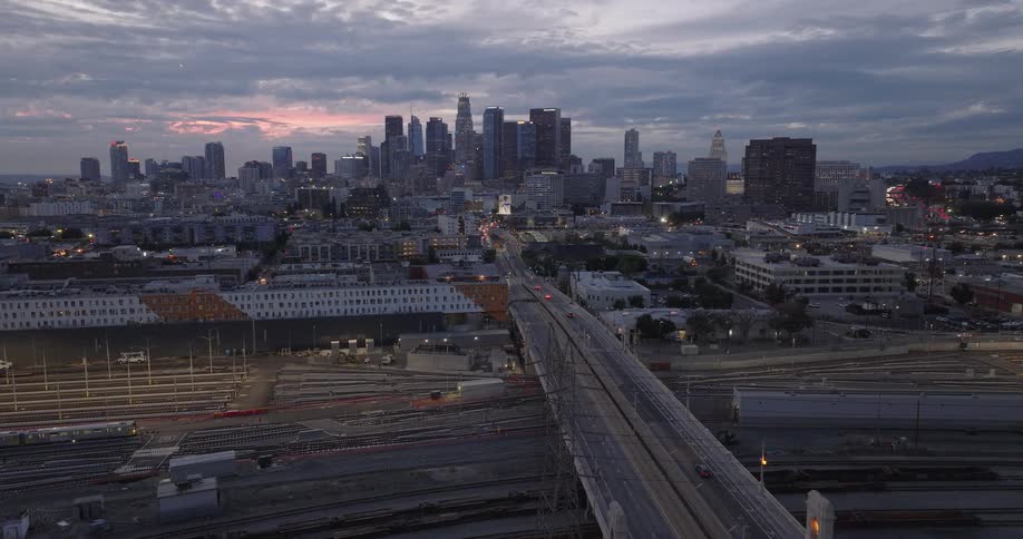 An aerial shot captures downtown Los Angeles skyline at dusk viewed over a wide railway yard and a central bridge with light traffic moving toward the illuminated buildings