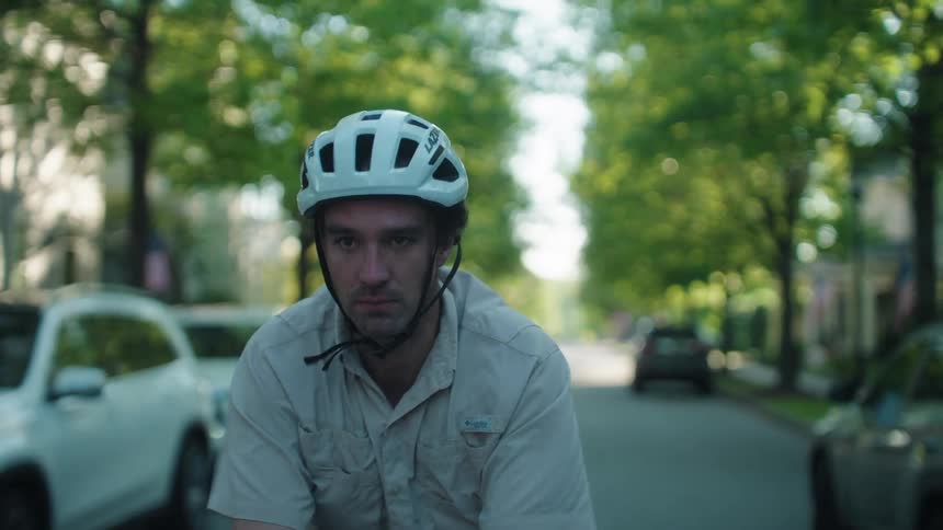 A man wearing a white bicycle helmet and a lightcolored collared shirt rides a bike down a shaded suburban street