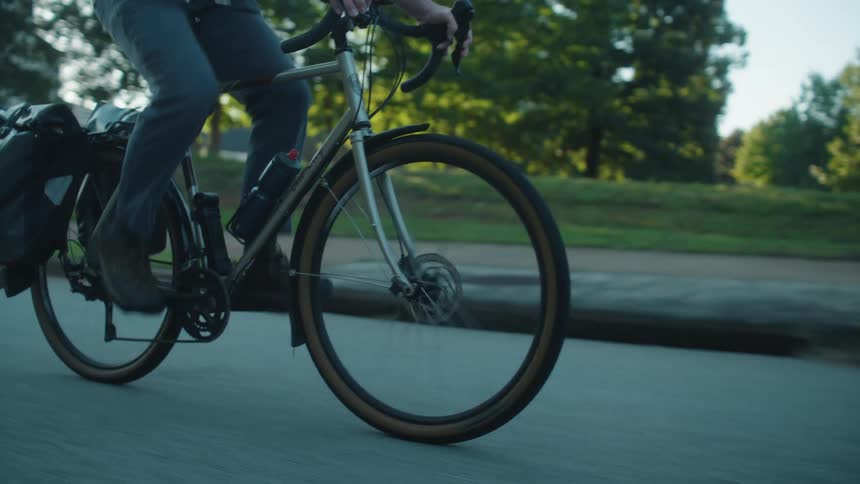 A person is shown riding a bicycle equipped with fenders and a cargo bag along a paved roadthe focus is on the lower half of the cyclist and the motion of the wheels with trees visible in the background against a dimming sky