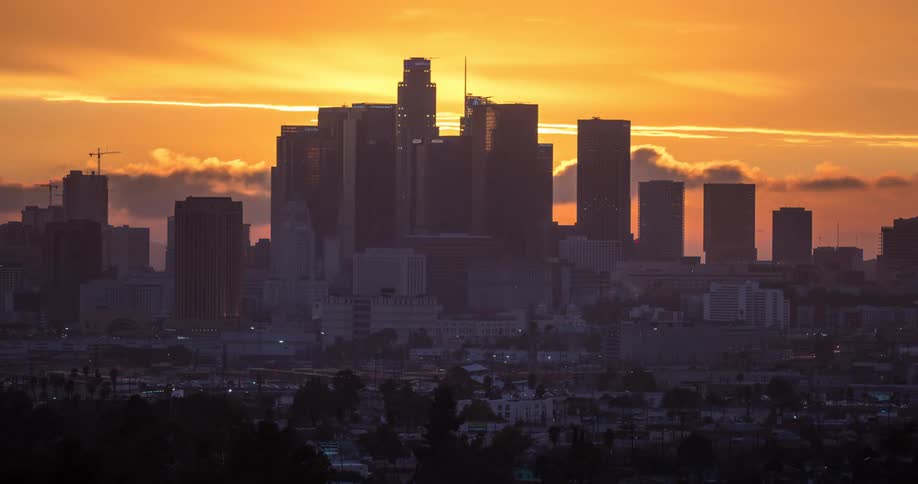 This is a stunning timelapse shot of a major city skyline likely downtown los angeles transitioning from sunset into twilight the sun dramatically sets behind the cluster of skyscrapers illuminating the clouds in brilliant shades of orange yellow a
