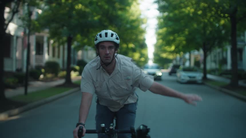 Man cycling through a residential neighborhood signals and turns as a car approaches behind him