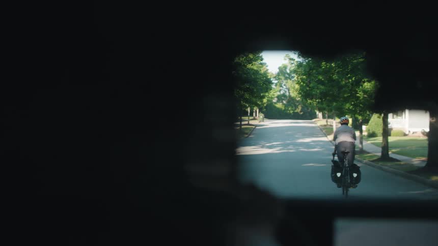 Man wearing a helmet and riding a motorized bicycle through a street on a suburban area making a sign for the driver of a car behind him before he crosses in a different direction during day time