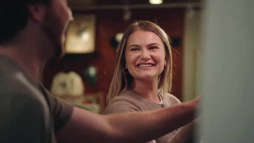 A young woman with a genuine happy smile is actively engaged in a lively conversation with a man whose shoulder is visible in the foreground in a warmly lit interior space