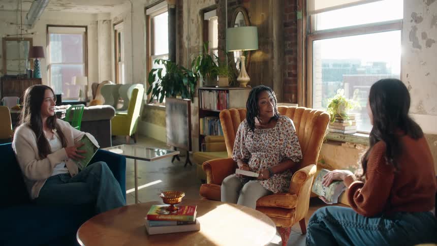 Three women are seated in a bright loftstyle room with large windows engaged in a conversation around a coffee table with one woman in the center smiling widely while holding a small book