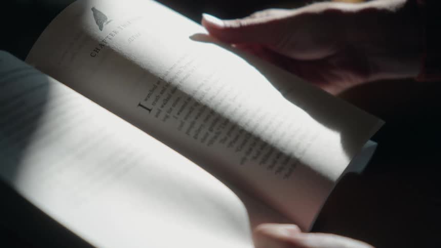 A persons hand partially visible in the dim light turns a page of an open book to reveal a new chapter title as a bright shaft of light illuminates the textured paper and printed text